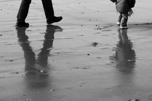 reflections at the beach: a man and a child walking at the beach, reflected in the wet sand.