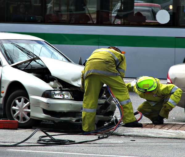 fire & rescue at work: firemen using jaws of life to open tangled car metal at accident scene with bus passing by