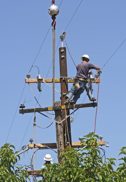 Men Replacing Power Pole: A crane drops a new power pole into a hole next to the one they are replacing. These workers transfer the hardware and hook up the wires to the new pole.