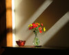 vase of flowers in shadows and: A vase of red and yellow flowers in rays of sun and shadow on a wooden table next to a ceramic bowl. Interesting how it looks as if a shadow is cast upon the flowers but they glow in the light anyway.